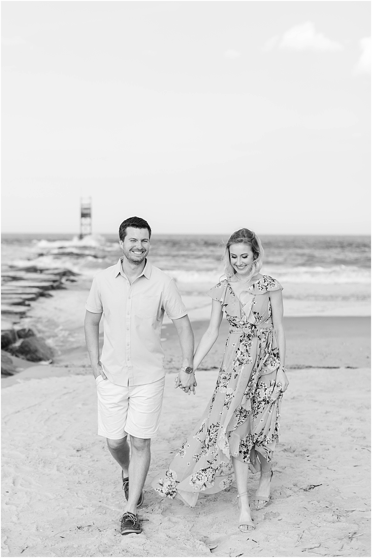 Black and white photo of Val and Don holding hands and smiling as they walk across Bethany Beach during their engagement session.