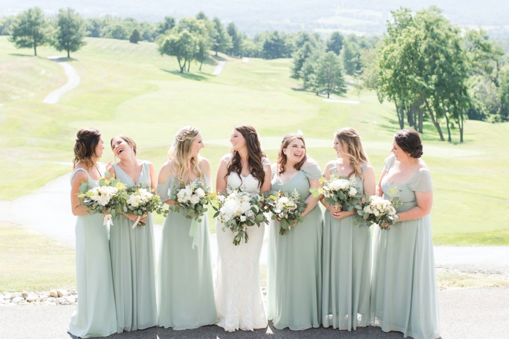 katie standing with her bridesmaids who are wearing sage green dresses and holding bouquets made of white flowers and greenery 