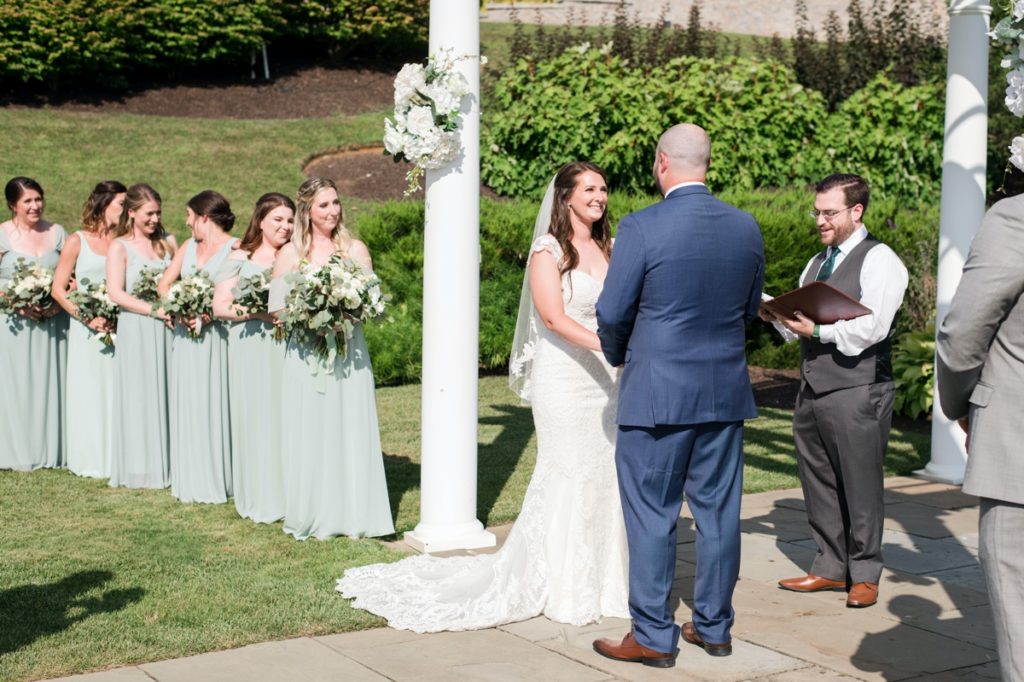 katie and welsey smiling at each other at the alter with the bridesmaids in the background during the ceremony at their Cotoctin Hall Wedding at the Musket Ridge Golf Club in Maryland 
