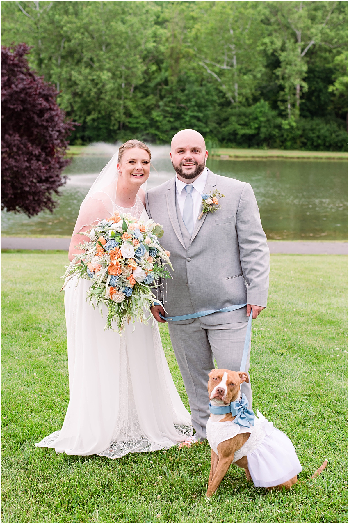 Anneliese and Alec smiling with their dog in front of a pond on their wedding day.